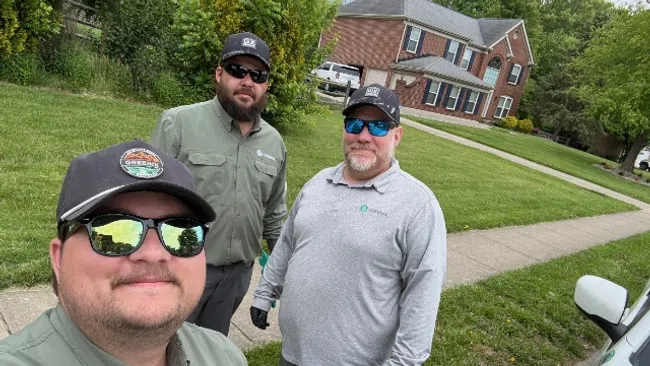 Three men wearing sunglasses and work uniforms posing outdoors with a residential house and sidewalk in the background.