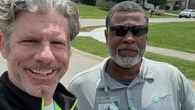 Two men outdoors on a sunny suburban street, one smiling and the other wearing sunglasses and blue gloves holding small items.