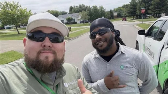 Two Greenix employees wearing branded shirts and sunglasses pose with thumbs up near a company vehicle on a suburban street.