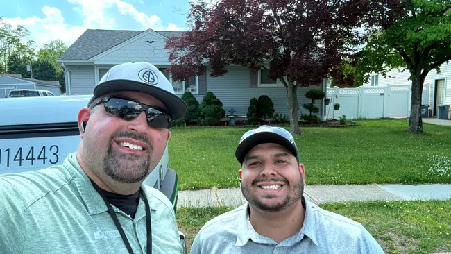 Two men wearing Greenix apparel smiling outdoors in front of a house and truck on a sunny day.