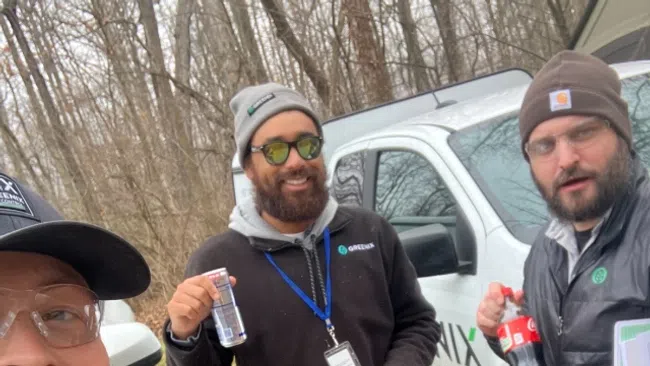 Three men wearing GRNIX apparel stand outdoors near company vehicles, holding drinks and smiling at the camera.
