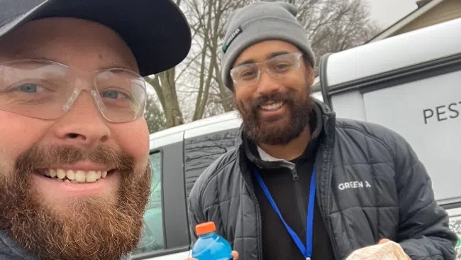 Two smiling pest control workers in branded jackets and badges stand outdoors by a company vehicle holding a drink and sandwich.