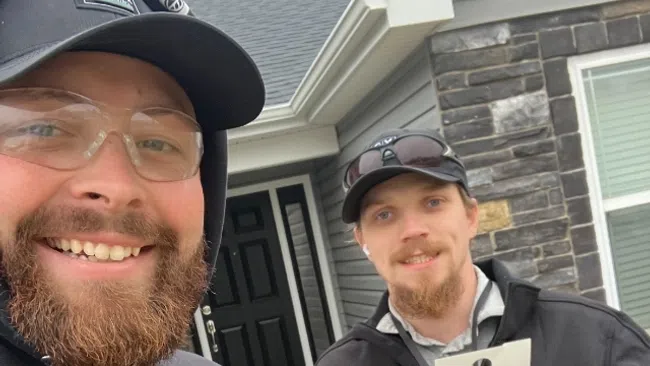 Two men in work attire smiling outdoors near a home entrance, one holding an informational flyer.