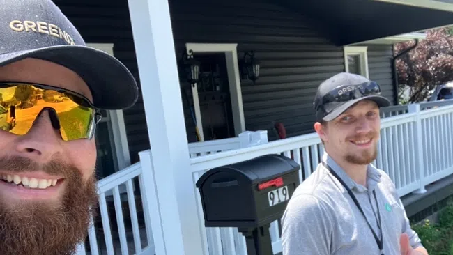 Two landscapers giving thumbs up outside a house with a porch and mailbox, one holding a red leaf blower.
