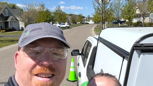 Two smiling pest control workers wearing safety glasses next to a branded service vehicle on a sunny suburban street.