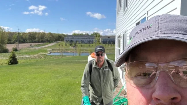Man in cap and safety glasses takes selfie while another man uses a backpack blower on green lawn next to white house.