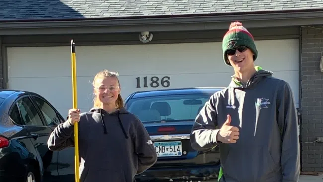 Two young people standing outside a garage, one holding a yellow broom, the other giving a thumbs up on a sunny day.