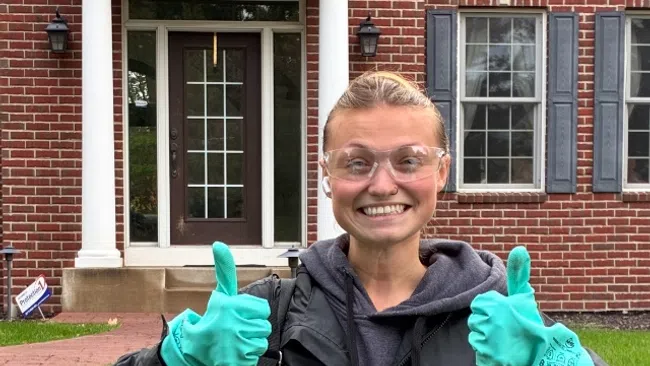 Smiling woman wearing safety glasses and teal gloves giving two thumbs up in front of a brick house.