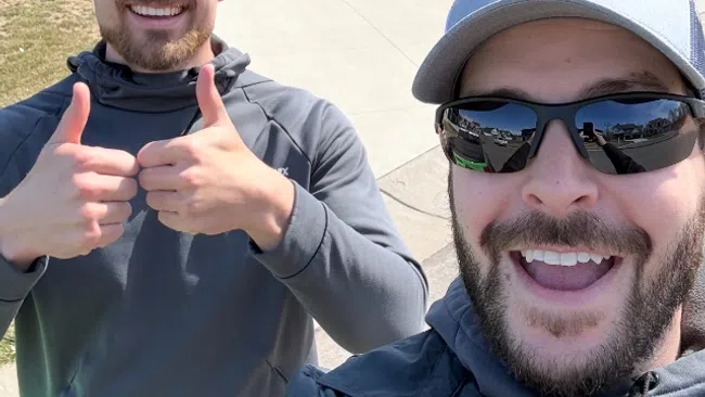 Two smiling men in sunglasses and hats give thumbs up outside a modern house with a three-car garage on a clear day