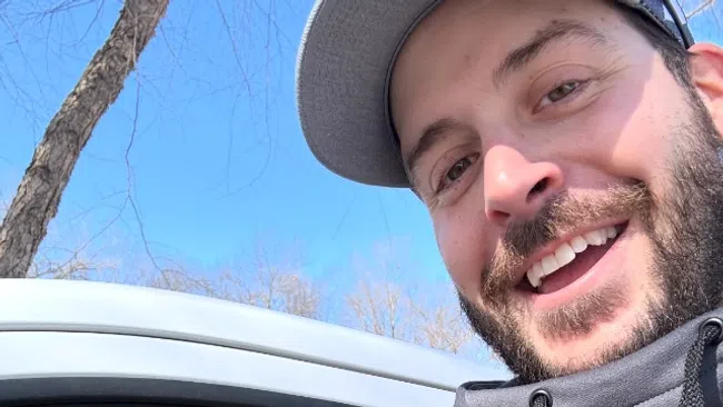Two smiling men posing for a selfie with one inside a car and clear blue sky in background