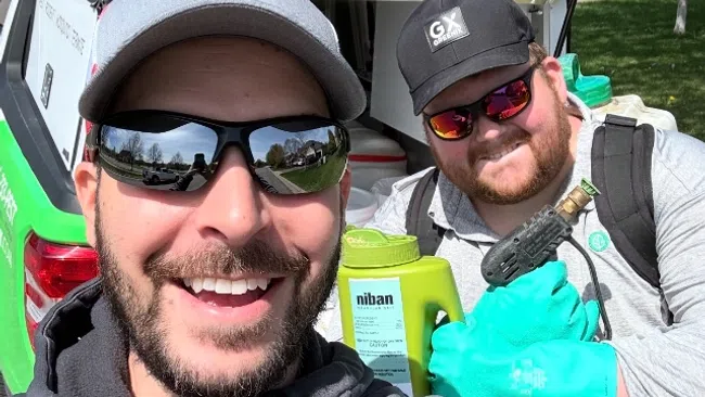 Two smiling pest control workers with sunglasses in front of a green and white service vehicle with equipment outdoors