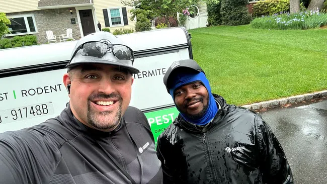 Two pest control workers wearing rain jackets and caps smiling outside a residential home with green lawn.