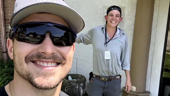 Two men smiling outdoors, one wearing sunglasses and cap, both dressed in casual work attire with Greenix logos visible.