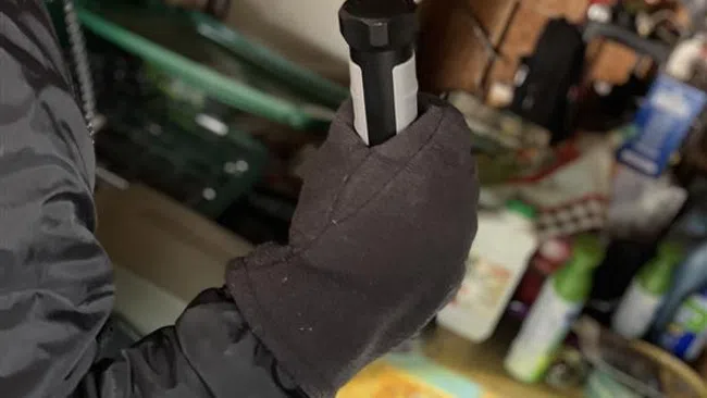 Man in gray beanie and gloves holding a black flashlight in a cluttered workshop or garage setting.