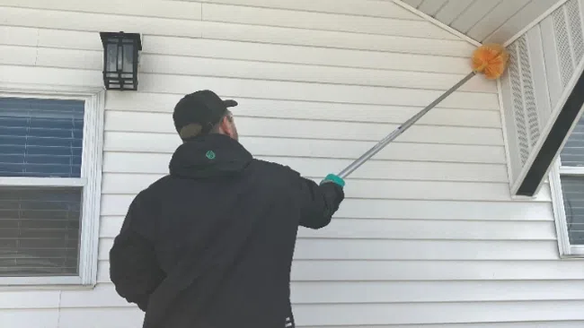 Person cleaning the house exterior soffit with a long-handled duster tool during daytime.