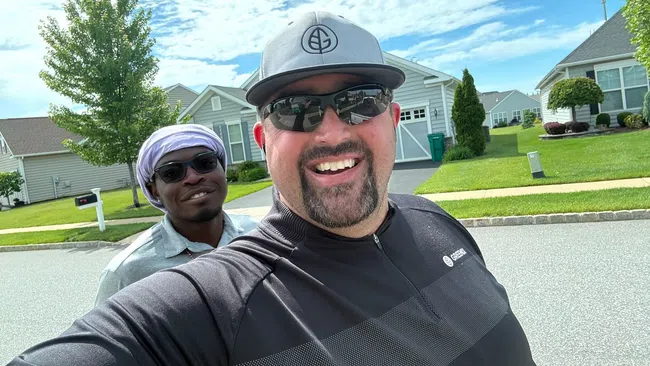 Two men smiling outdoors in a suburban neighborhood on a sunny day with blue sky and houses in background