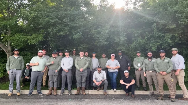 Group photo of 18 outdoor workers wearing uniforms standing and sitting in front of dense green trees with sunlight streaming through.