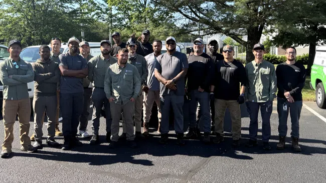 Group of utility workers standing in parking lot with work trucks and trees in the background on a sunny day