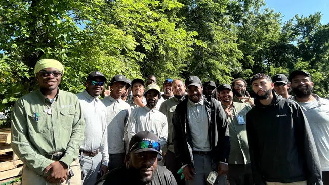 Group of diverse men posing outdoors in casual workwear under green trees on a sunny day