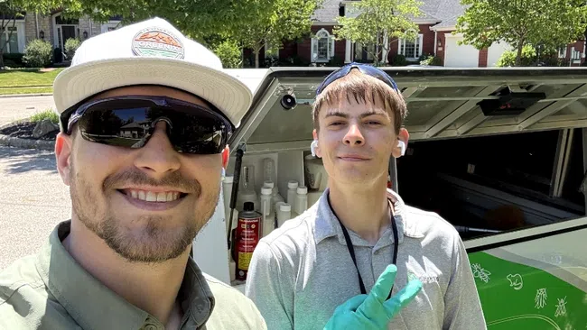 Two pest control workers smiling outdoors in front of a service truck with equipment on a sunny day.