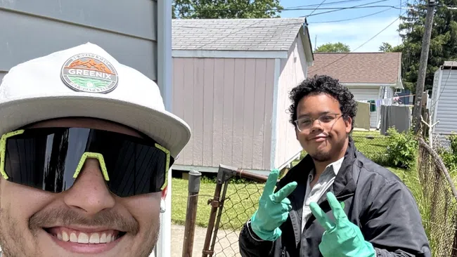 Two men outdoors smiling, one wearing a Greenix cap and shirt, another making peace signs with green gloves on a sunny day.