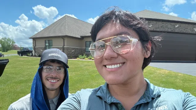 Two smiling men in Greenix uniforms outdoors on a sunny day with houses and green lawn in the background