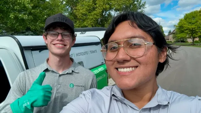Two pest control workers smiling in uniform by a company van on a sunny suburban street.