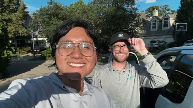 Two men smiling outdoors near a white vehicle in a residential neighborhood on a sunny day.