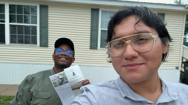 Two smiling people taking a selfie outdoors in front of a house, one holding a flyer or brochure.