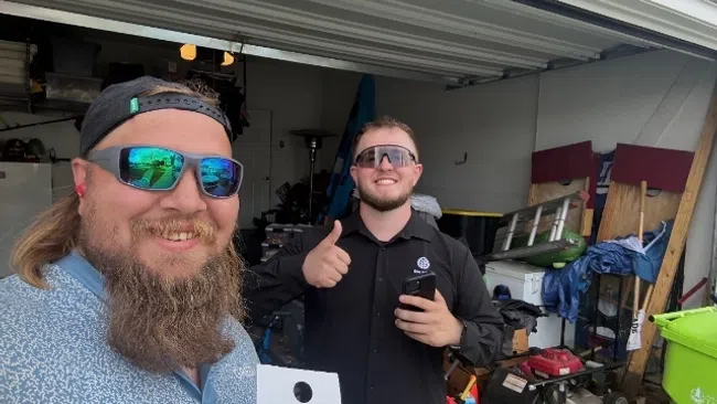 Two men wearing sunglasses smiling in a garage, one holding a Pest Nerds sign and the other giving a thumbs up.