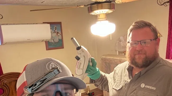 Two pest control workers in protective gear inside a room, one holding a spray device and both smiling at the camera.