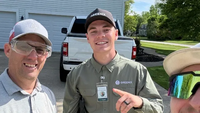Three Greenix workers in uniform smiling outdoors near a white garage and pickup truck on a sunny day.