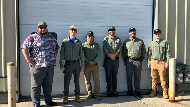 Six men in work uniforms standing outside a large industrial building in front of a closed white garage door.