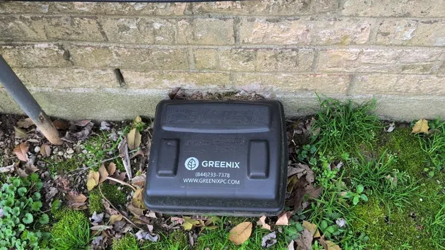 Black Greenix irrigation control box on mossy ground against a brick wall with overhead wiring.