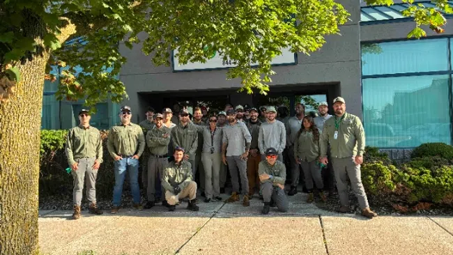 Group of uniformed workers posing outside a building on a sunny day with greenery and a tree in front