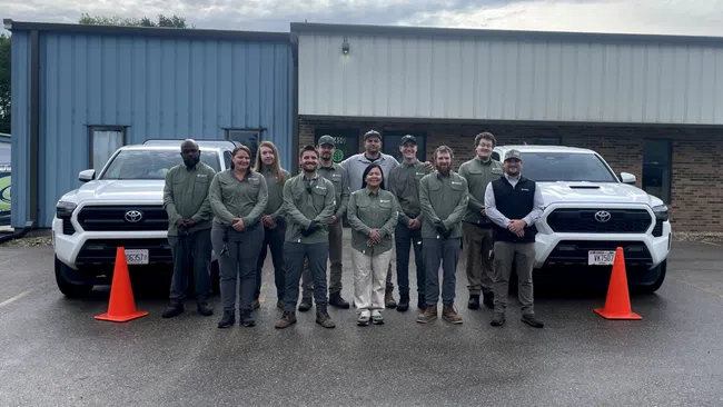 Team of eleven people in matching uniforms standing between two white Toyota trucks outside a commercial building.