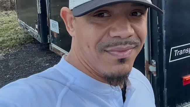 Man wearing GRNX hat and shirt taking selfie in front of a black Haulmark trailer outdoors.