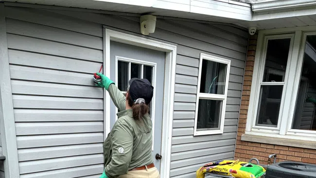 Person in green jacket and gloves cleaning exterior gray siding of a house near a white door and garden supplies.