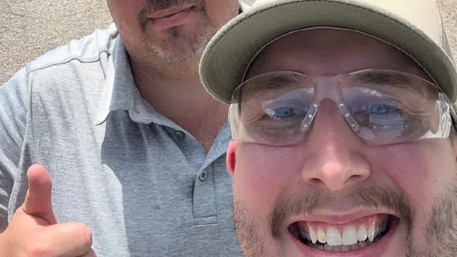 Two men wearing hats and sunglasses smiling outdoors on a sunny day with green trees and blue sky behind them.
