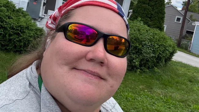 Person wearing sunglasses and a striped bandana outdoors with greenery and houses in the background