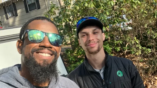 Two men outdoors smiling, one with sunglasses and beard, the other wearing a cap giving a thumbs up in a wooded area.