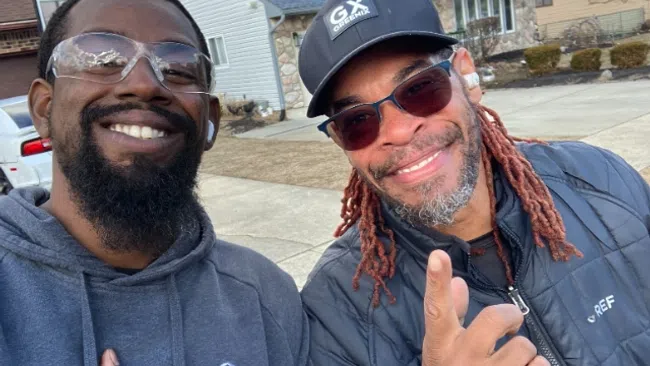 Two men smiling outdoors, one wearing a GREEFIX hoodie and the other a black jacket and cap with sunglasses.