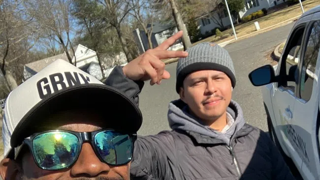 Two men smiling outdoors on a sunny day, one wearing sunglasses and a cap, the other wearing a beanie and jacket.