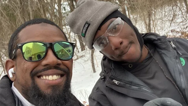 Two men in winter jackets and gloves smiling outdoors in a snowy forest with leafless trees behind them