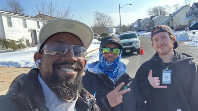 Three men wearing jackets and hats posing outdoors on a sunny winter day in a suburban neighborhood.
