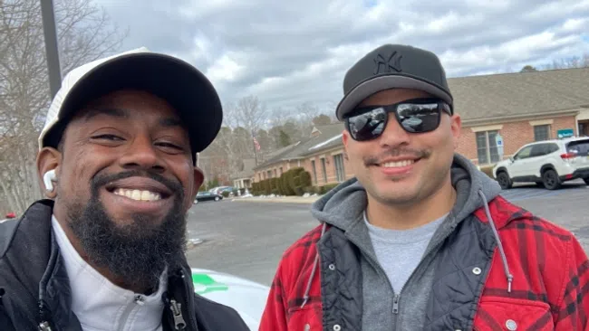 Two smiling men outdoors wearing jackets and caps with buildings and cars in the background on a cloudy day