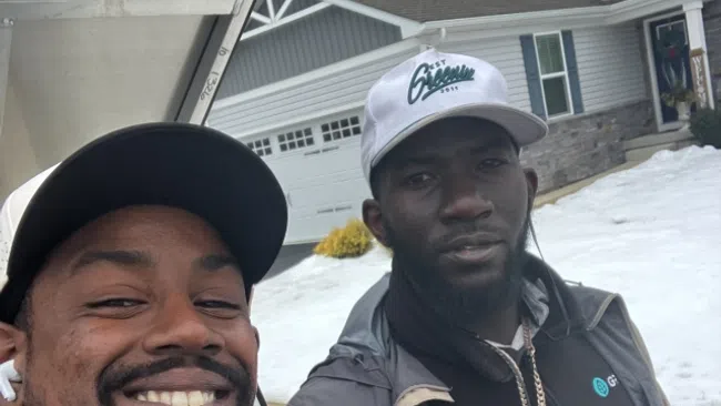 Two men outdoors in winter clothing, one smiling and the other giving a thumbs-up in front of a house with snow.