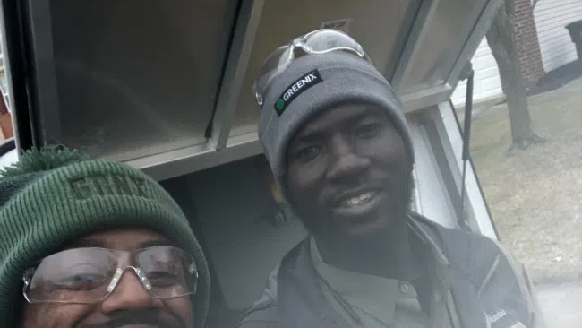 Two smiling men wearing outdoor work jackets and beanies standing near an open truck door in a residential area.