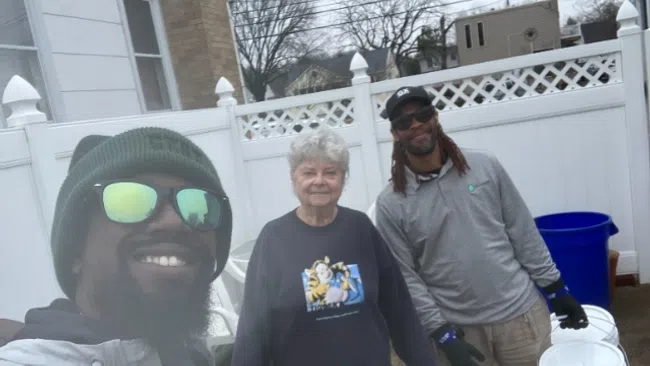 Two men and an elderly woman smiling outdoors near white fence holding buckets on cloudy day