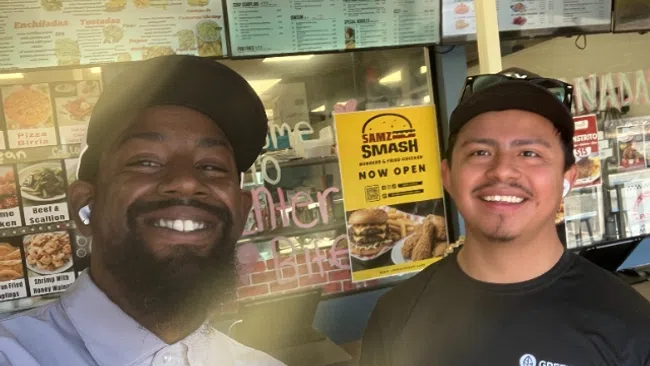 Two smiling men wearing caps pose inside a casual restaurant with colorful menu boards behind them.